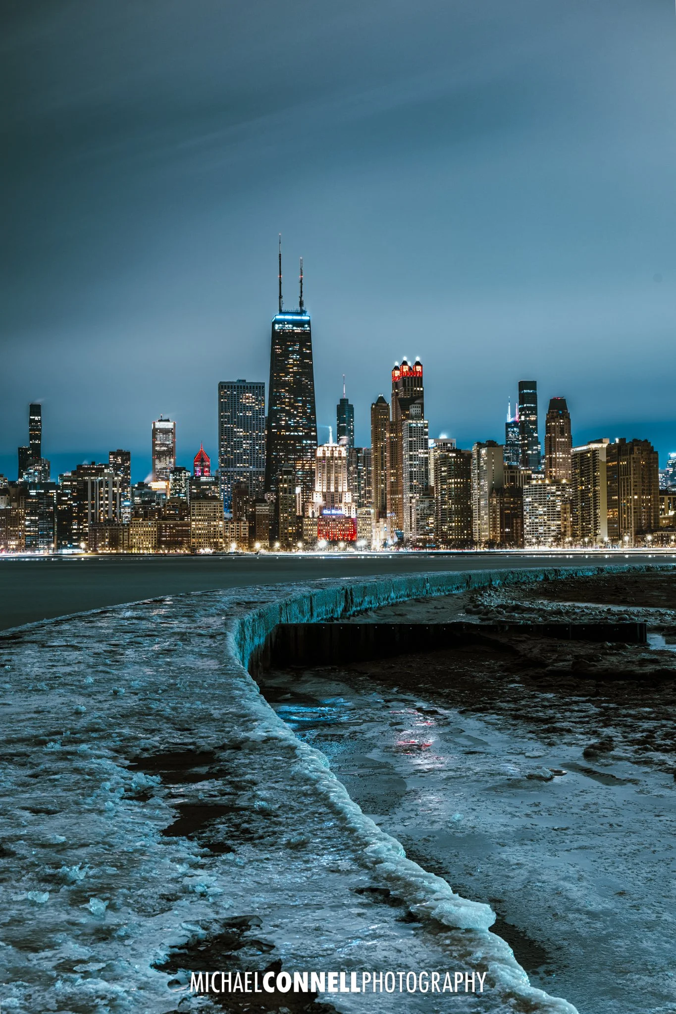 Nighttime cityscape of Chicago skyline viewed from the waterfront, with prominent tall buildings and a cloudy sky.