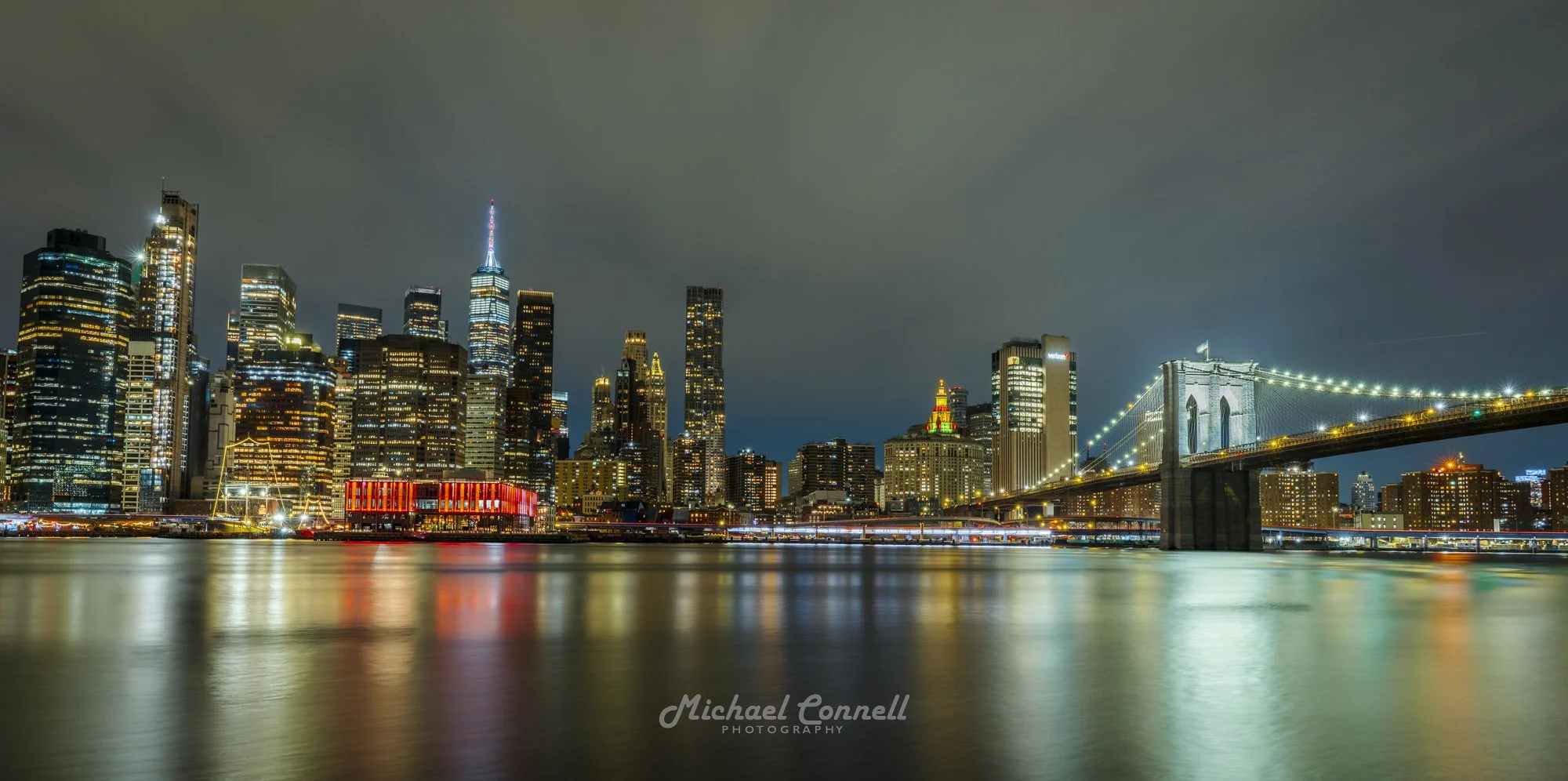 Nighttime view of Manhattan skyline with illuminated skyscrapers and Brooklyn Bridge reflecting on the water.
