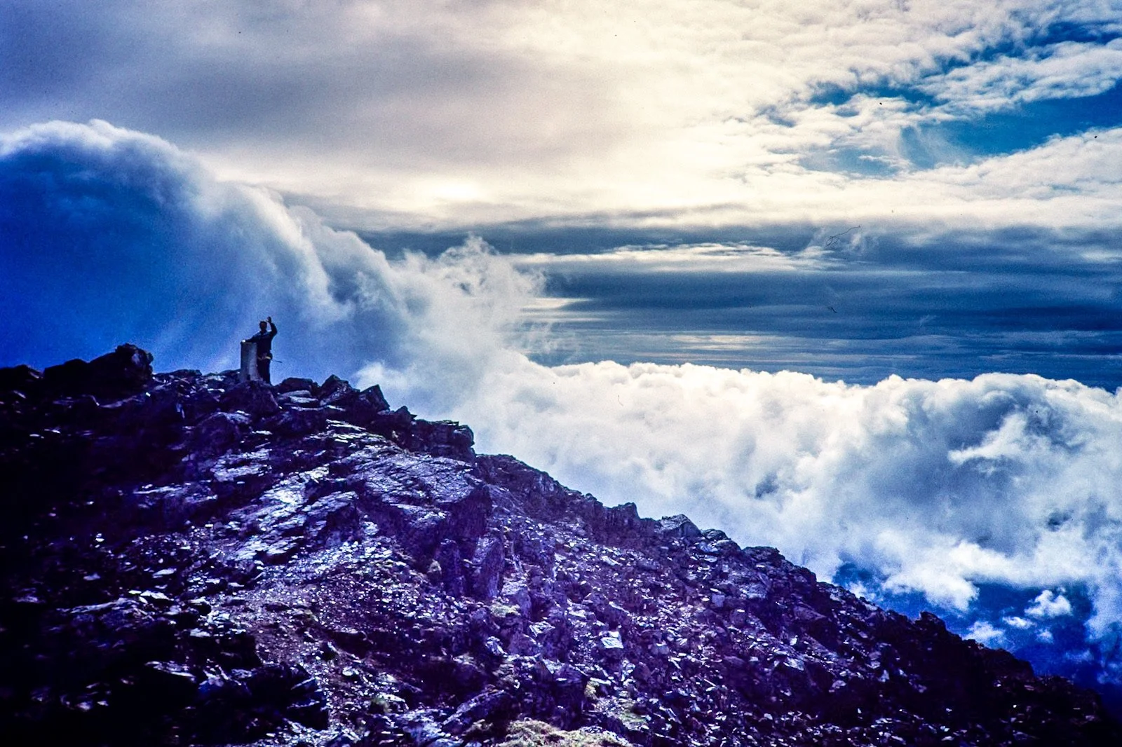 Person standing on rocky mountain peak with clouds and sky in the background, some clouds are dark and stormy, others are light and wispy.