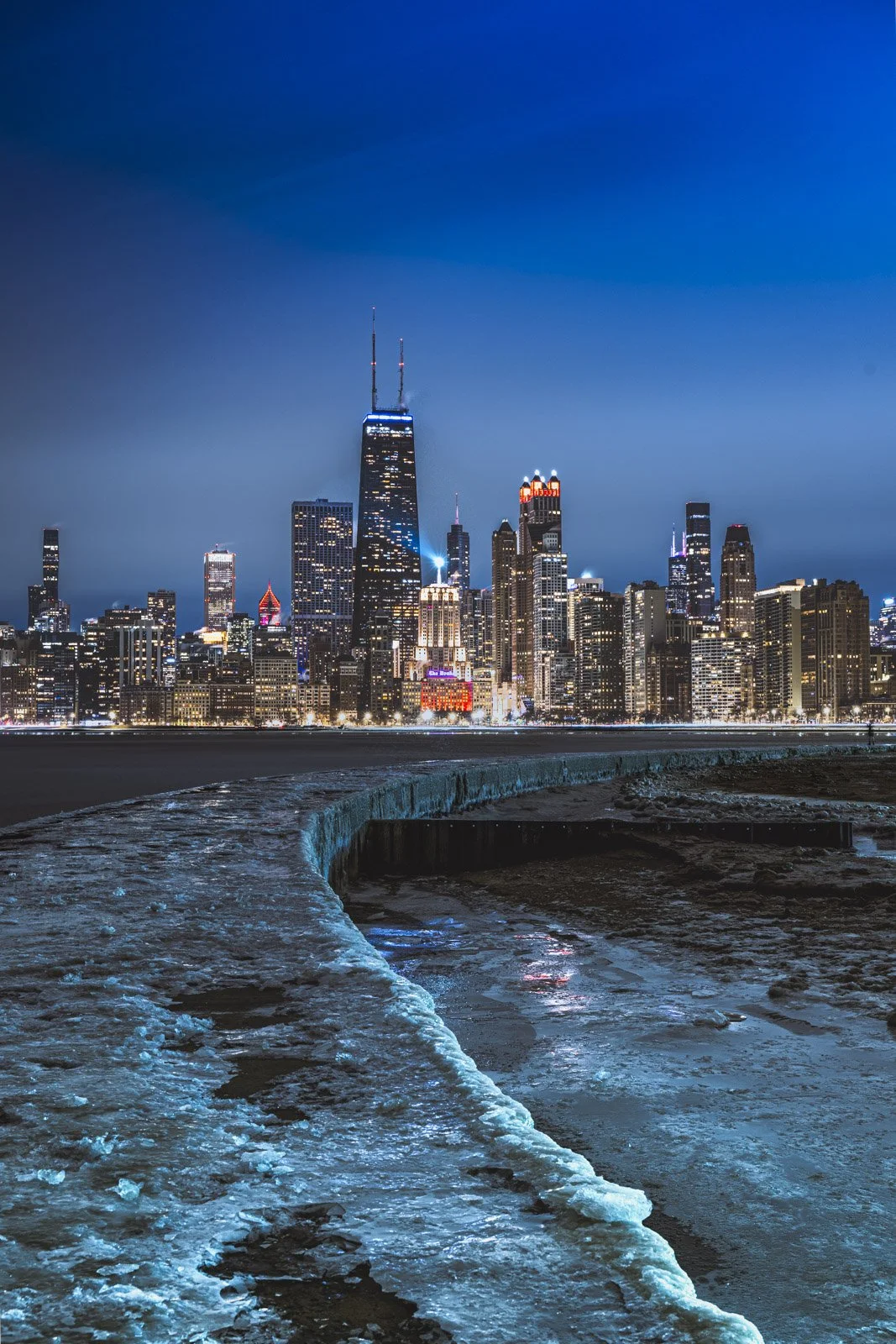 Nighttime city skyline of downtown Chicago with tall buildings illuminated, viewed from icy waterfront with frozen patches and snow in the foreground.