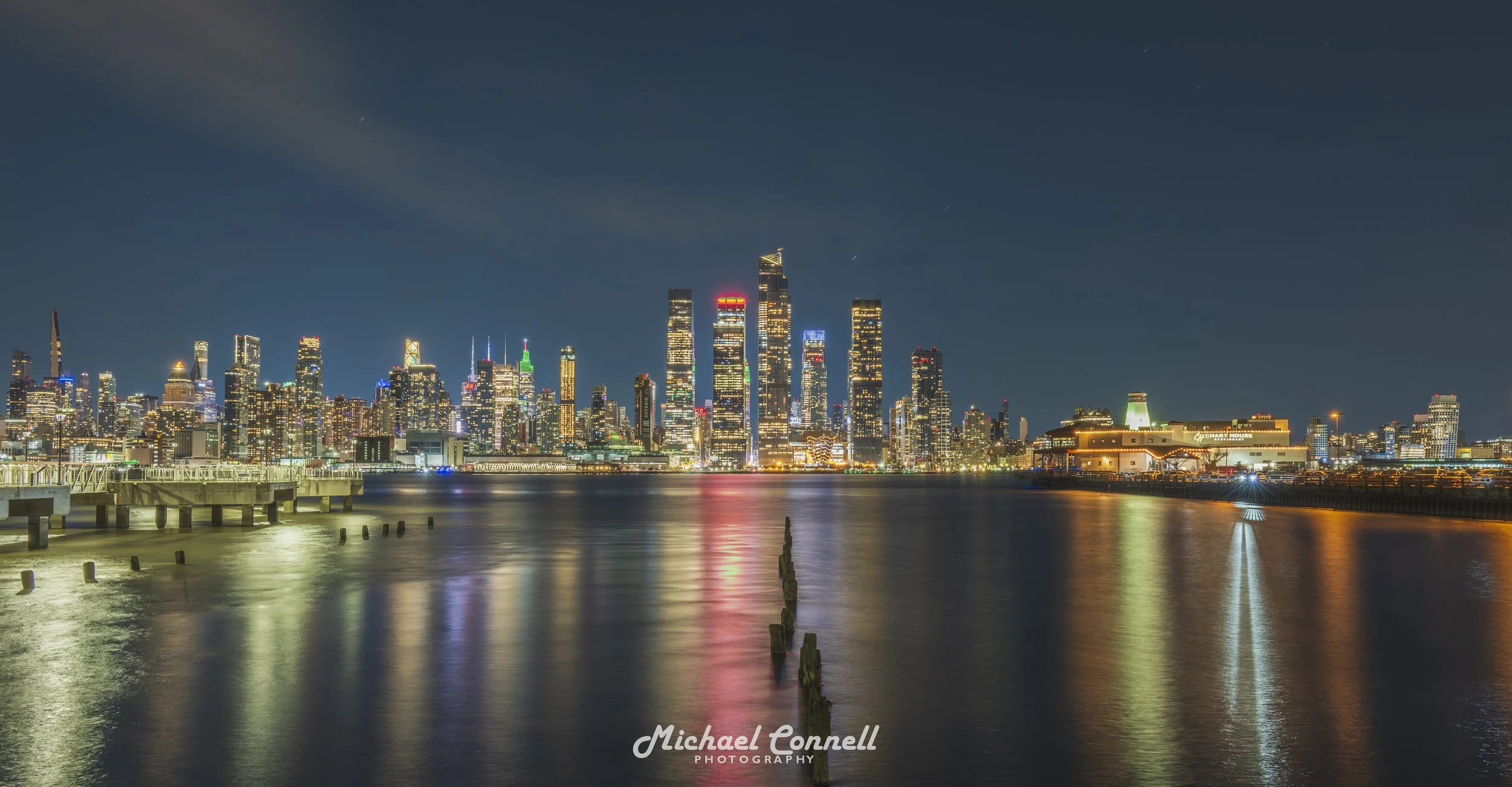 Nighttime view of a city skyline with illuminated skyscrapers reflecting on a body of water in the foreground, and a pier on the left side.