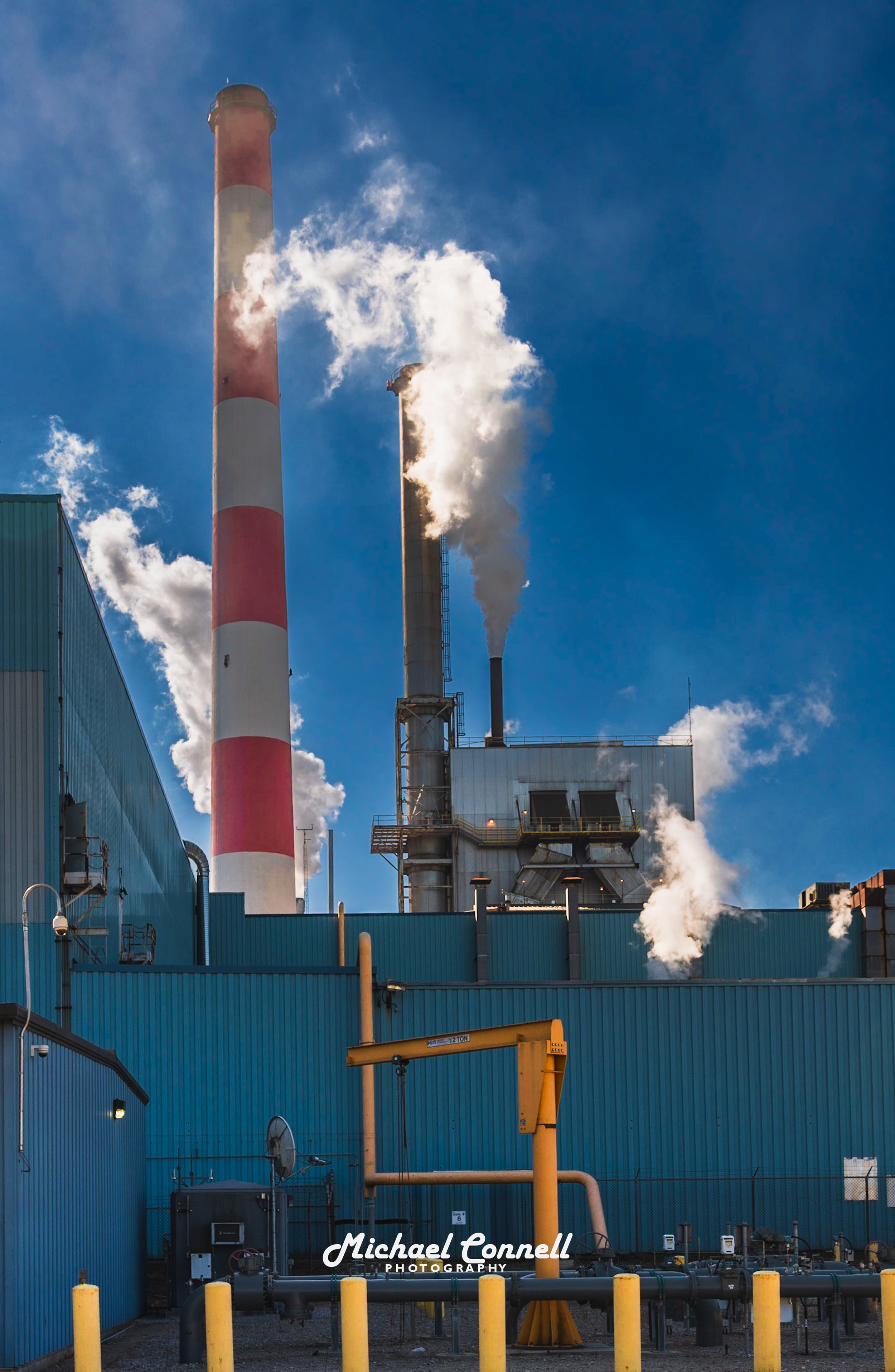 Industrial factory with blue buildings and multiple smokestacks emitting white smoke against a blue sky.