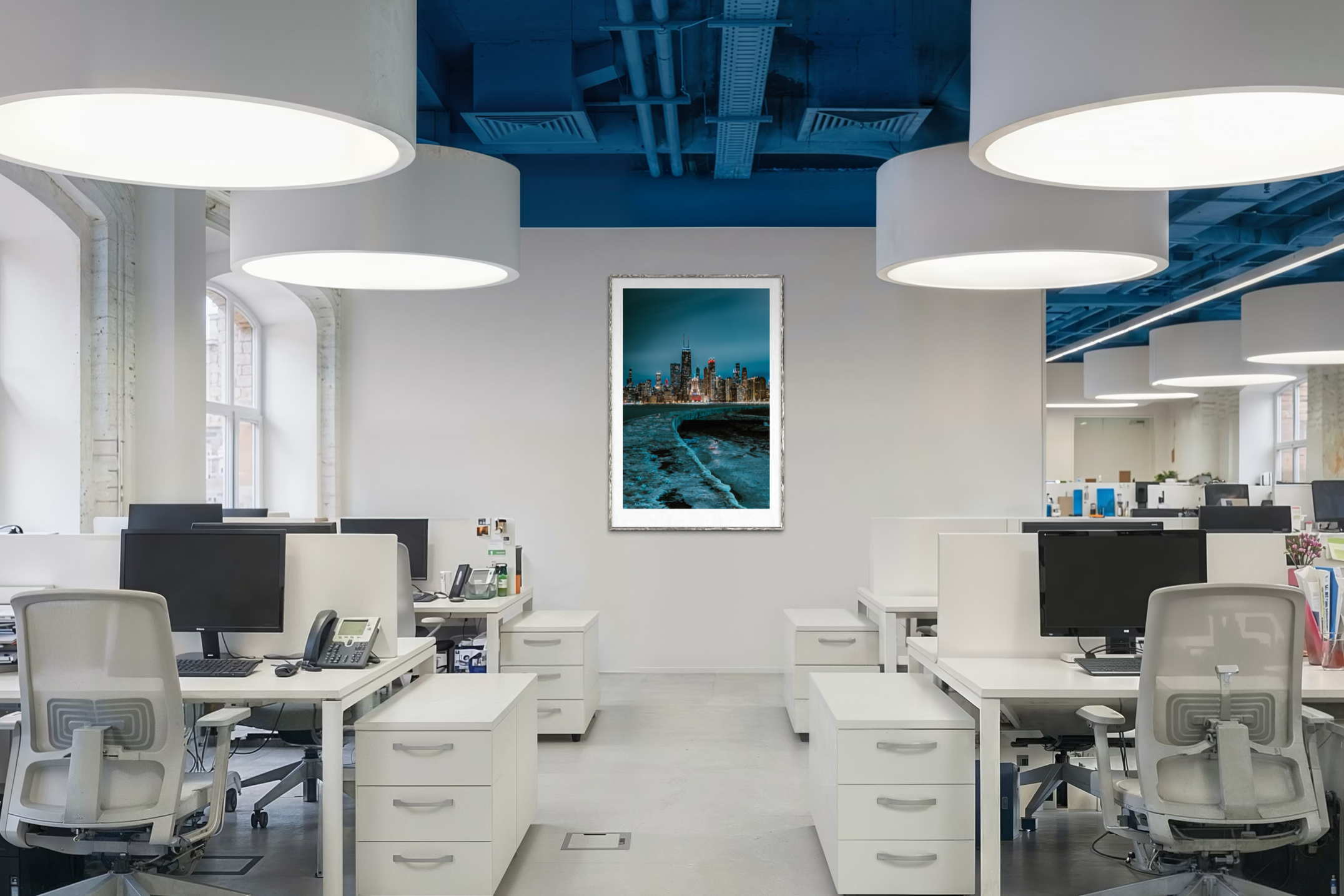 An open-plan office with white desks, office chairs, and computers. Large windows let in natural light. A framed cityscape photograph is on the white wall near the ceiling, with a blue ceiling and modern light fixtures.