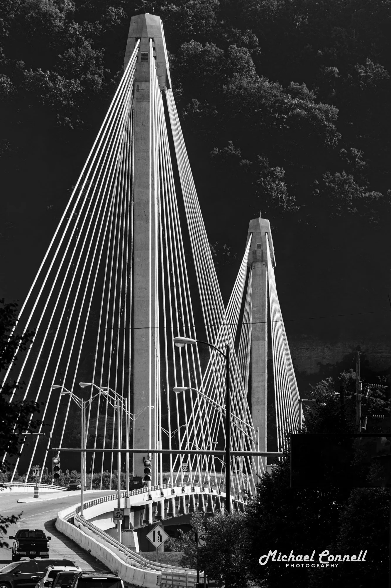 Black-and-white photo of a modern suspension bridge with tall towers and numerous cables, spanning over a road with cars and streetlights underneath.