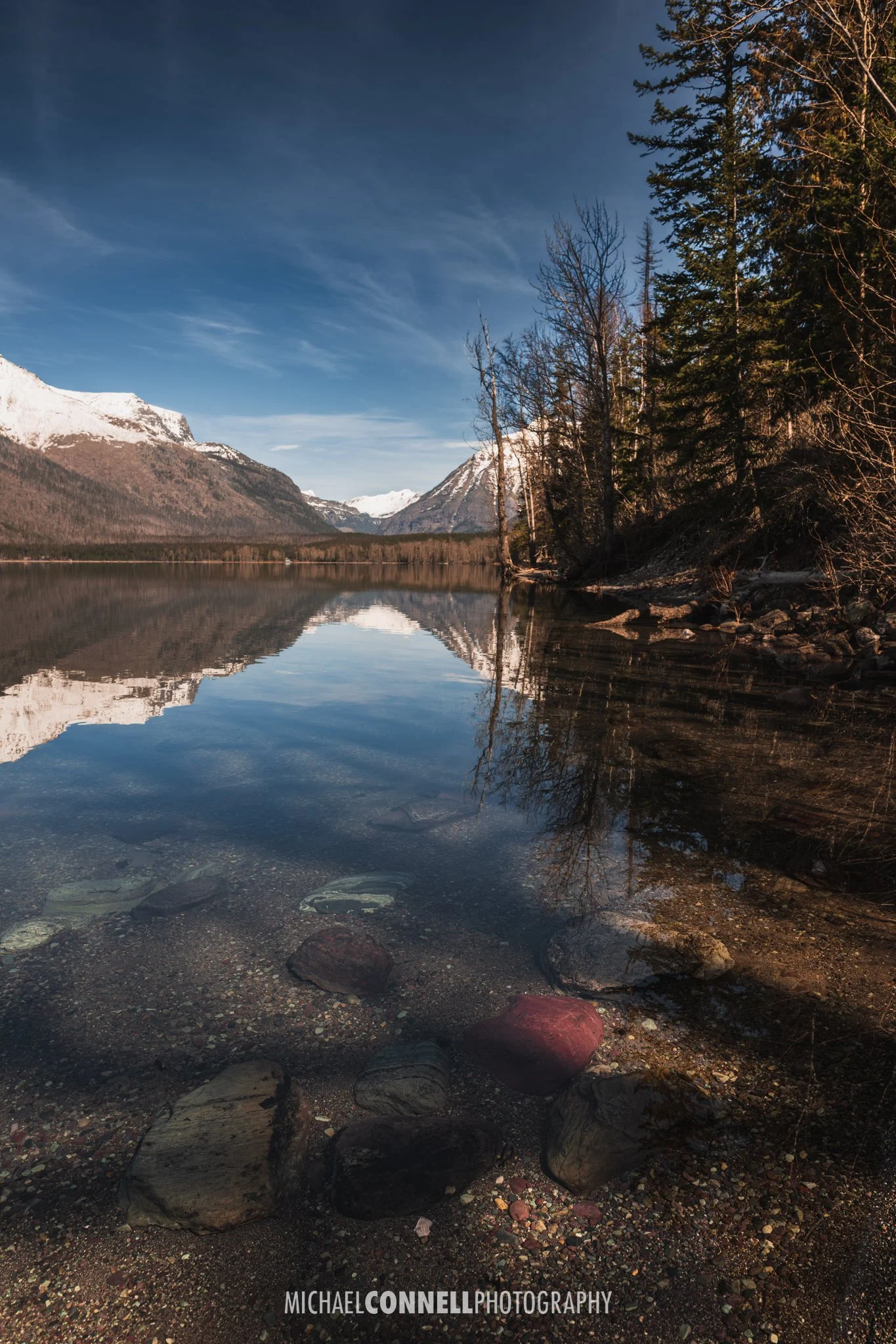 Scenic mountain lake with snow-capped peaks, clear reflective water, and a rocky shoreline lined with trees.
