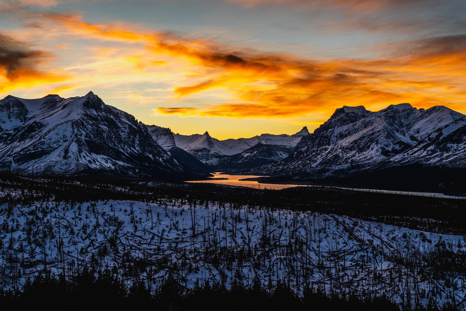 Snow-covered mountains at sunset with a river reflecting the sky and a partly cloudy orange and yellow sky.