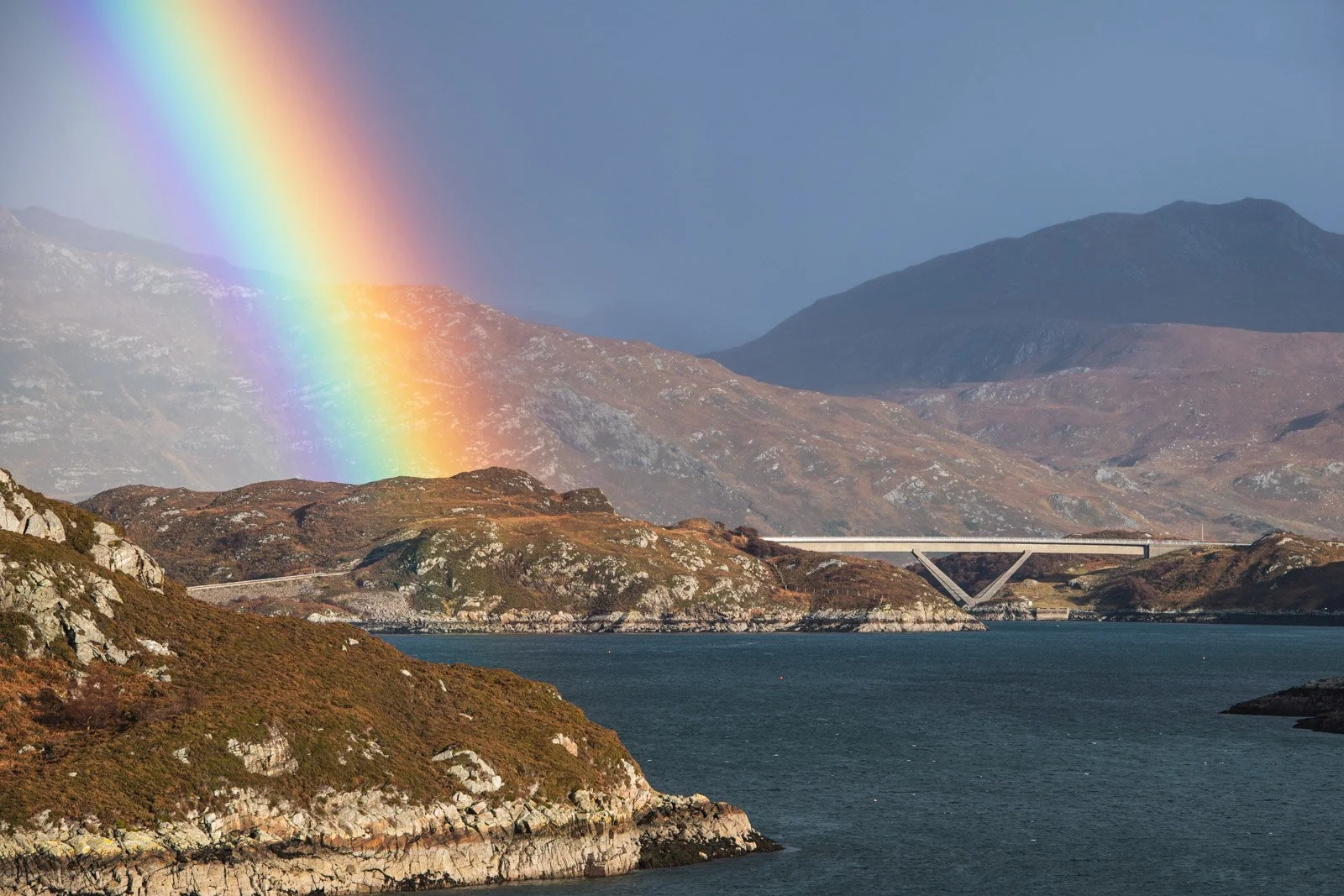 A scenic landscape featuring a rainbow arcing over mountainous terrain with a body of water in the foreground, and a bridge crossing the water in the distance.