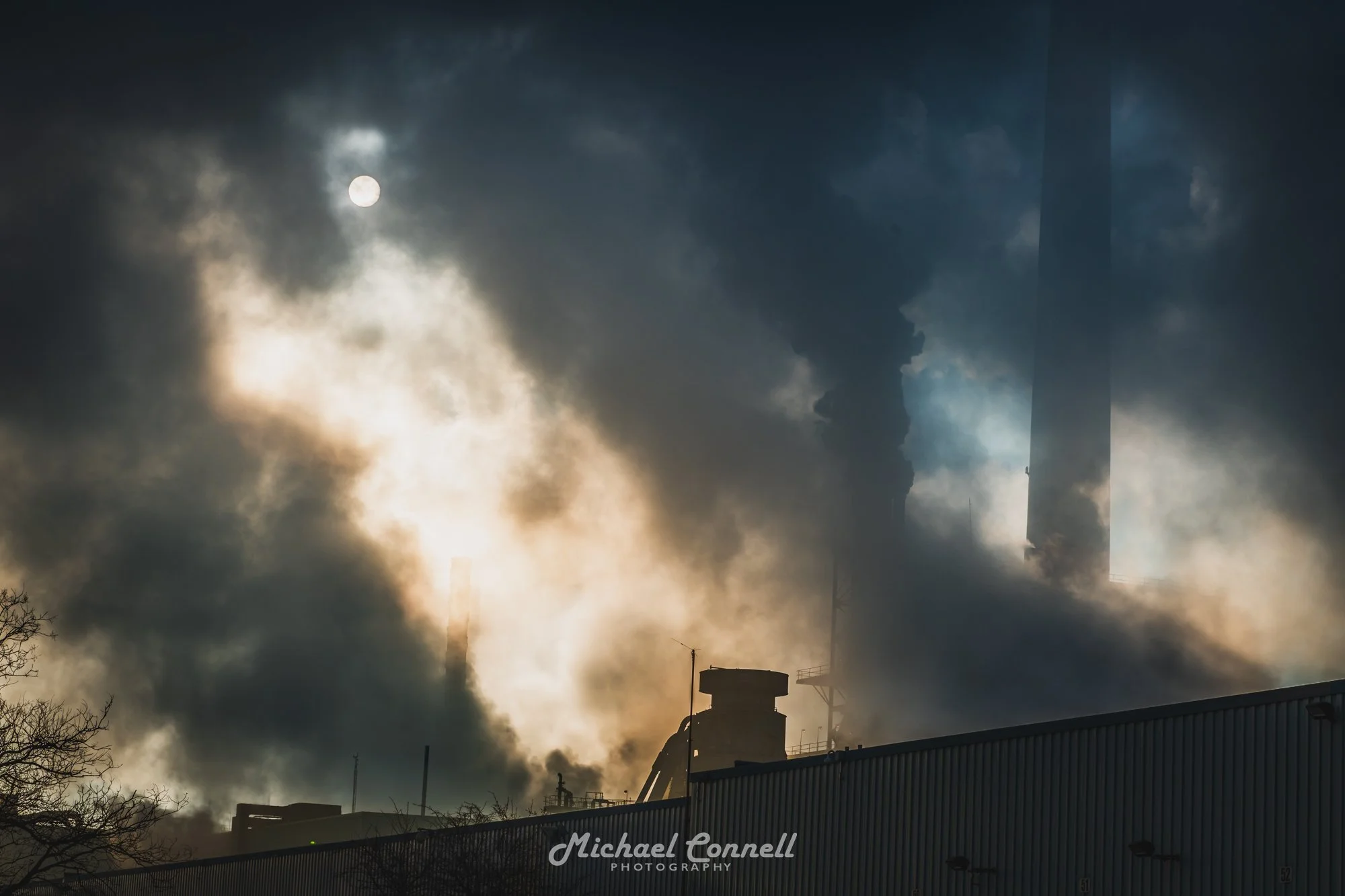 Industrial factory with smokestacks emitting thick black smoke against a dark sky, with the sun partially visible through clouds.