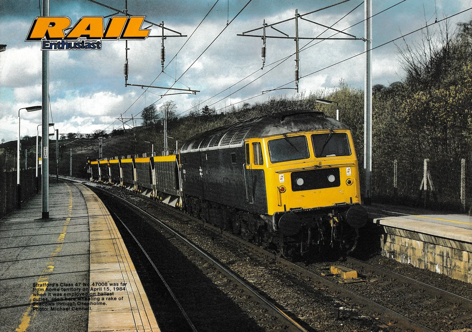A yellow and black class 47 train on the tracks at Oxenholme station with overhead electric lines and a cloudy sky in the background. The station platform has yellow safety markings and some signage visible. Rear cover of June 1985 "Rail Enthusiast"