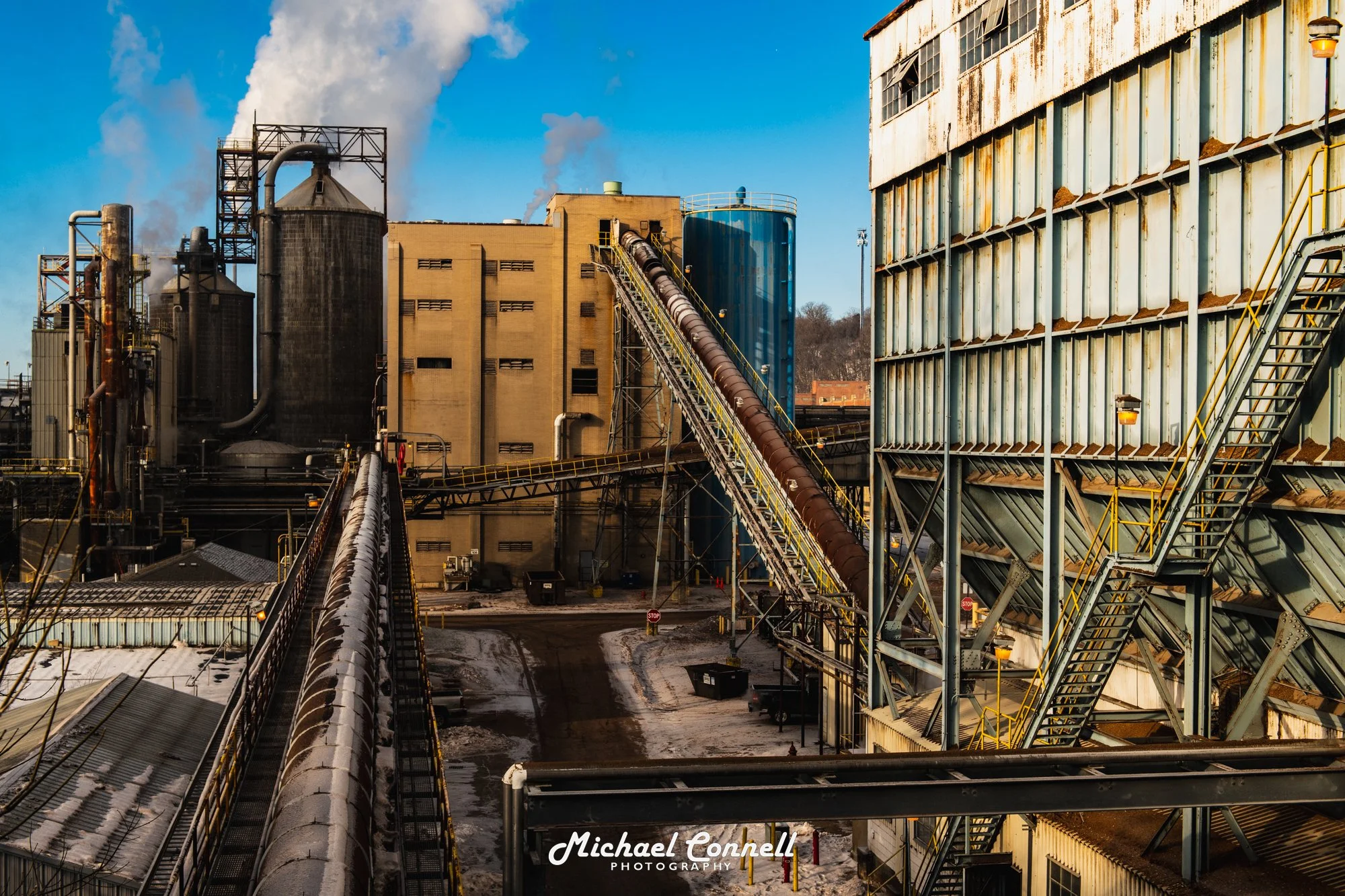 Industrial factory with large buildings, silos, and conveyor belts in a snowy environment, under a partly cloudy sky.