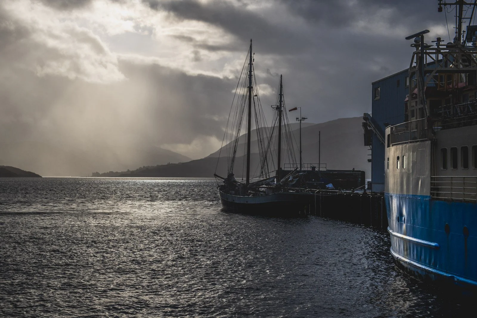 A harbor scene at sunset with cloudy sky, a sailboat docked next to a large blue boat, and mountains in the background.