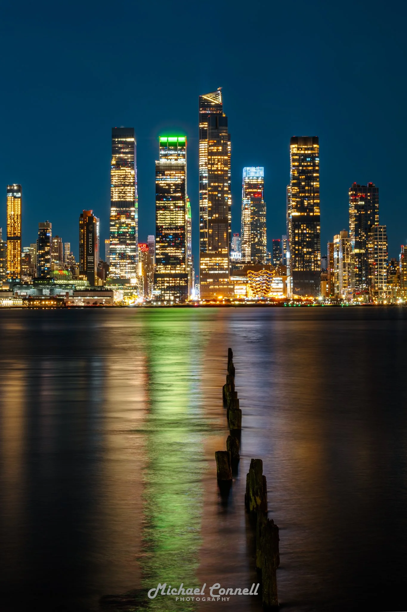 Nighttime view of a city skyline with illuminated skyscrapers and reflections on water, with old wooden posts in the foreground.