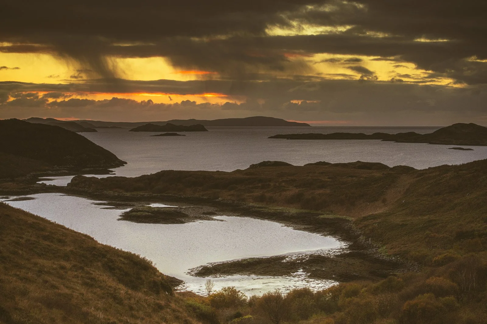 A scenic view of a river winding through green hills with a large body of water in the background, under a dramatic cloudy sky at sunset.