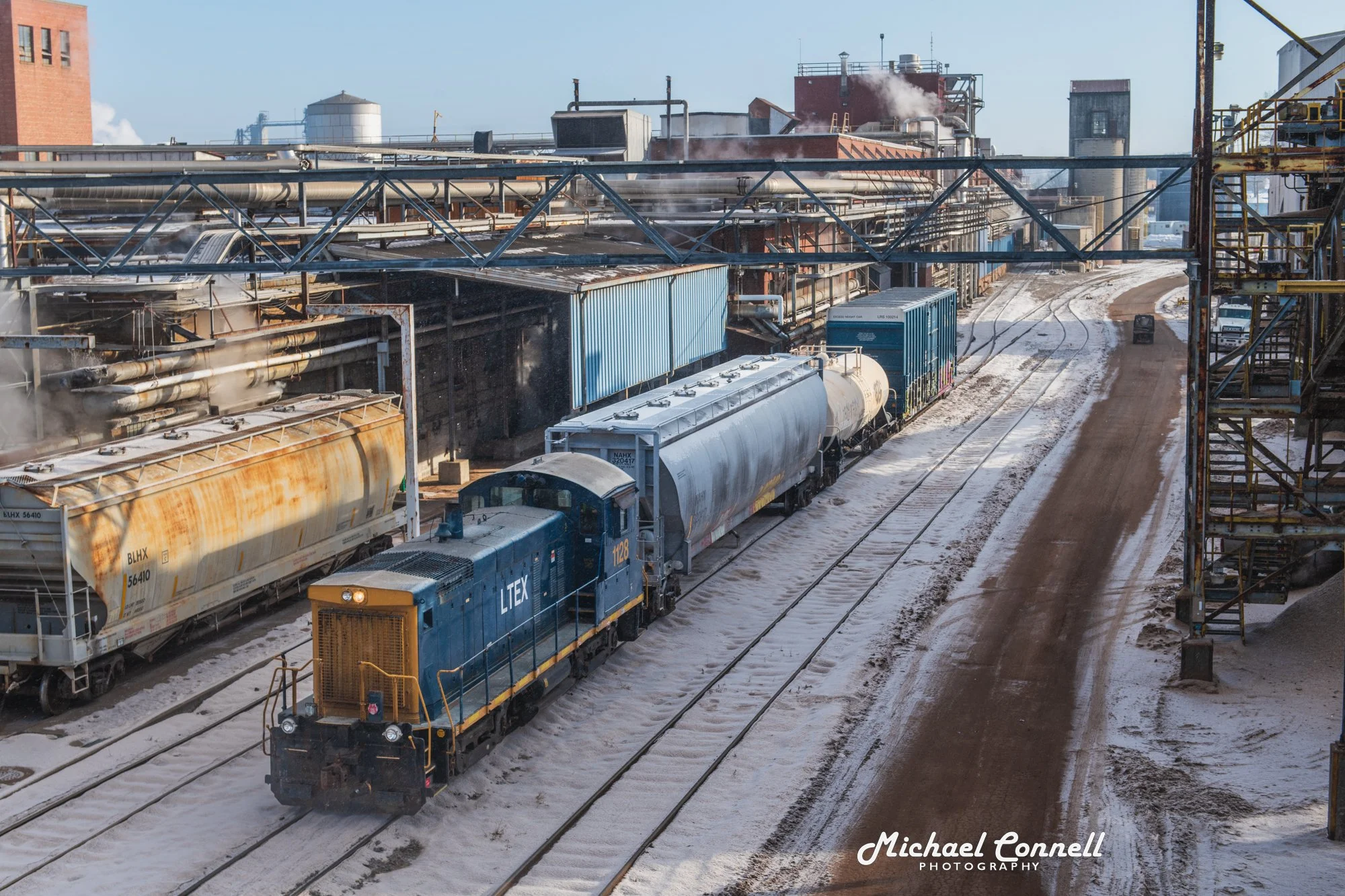 A blue locomotive train traveling on snow-covered railway tracks at an industrial site with storage tanks, pipes, and structures in the background.