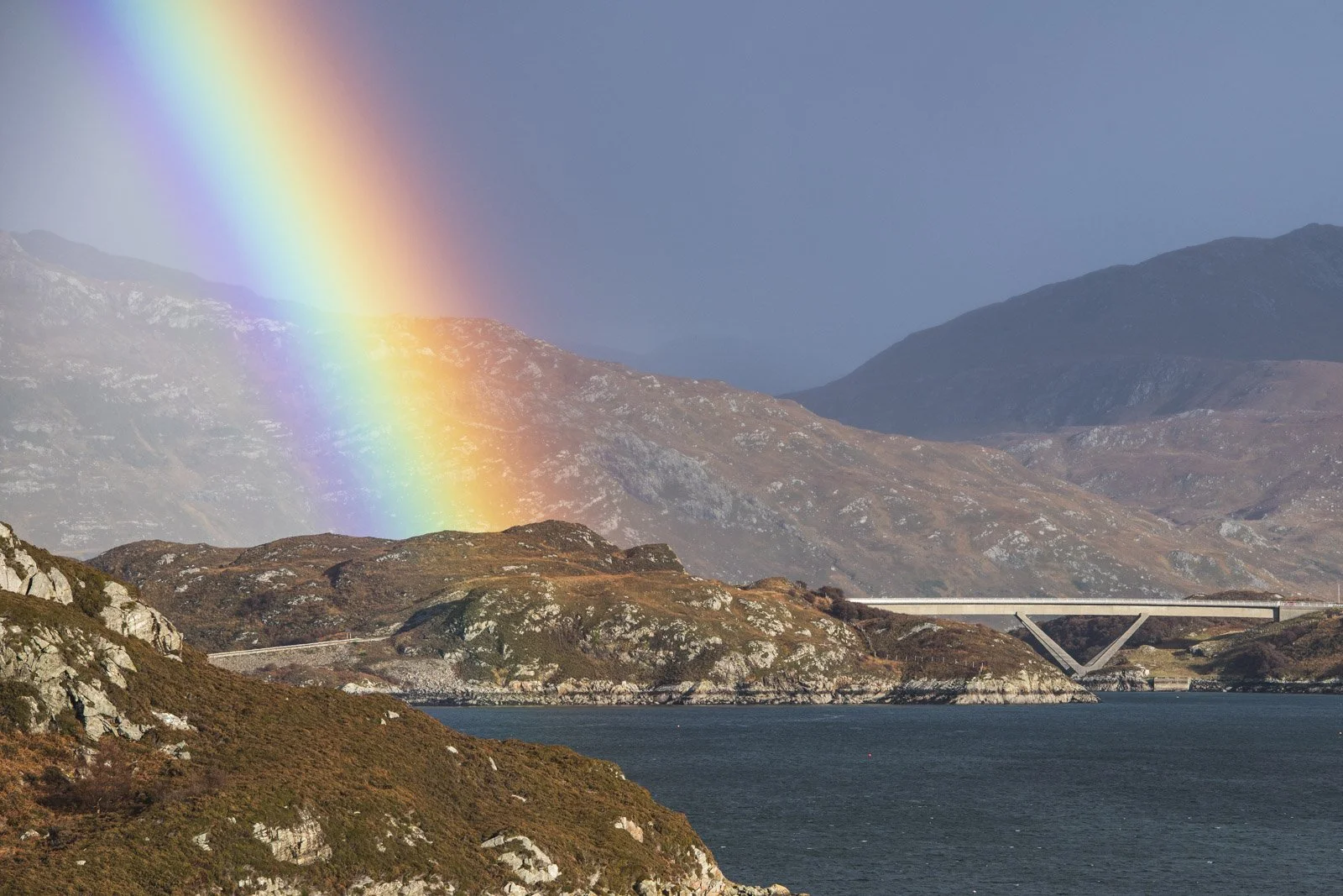 A rainbow arching over a mountainous landscape with a bridge spanning a body of water.