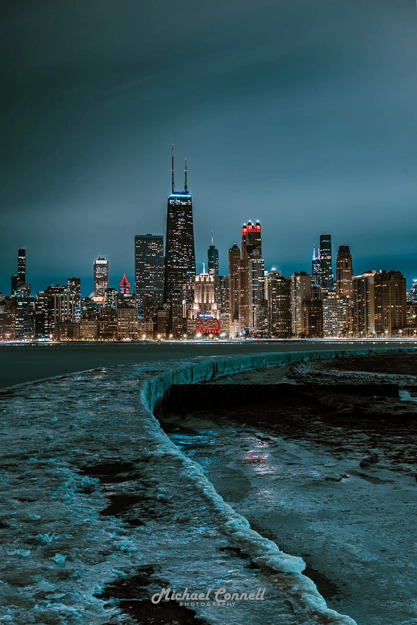 Nighttime view of downtown Chicago skyline with illuminated skyscrapers seen from a frozen shoreline with a curved ice-covered edge in the foreground.