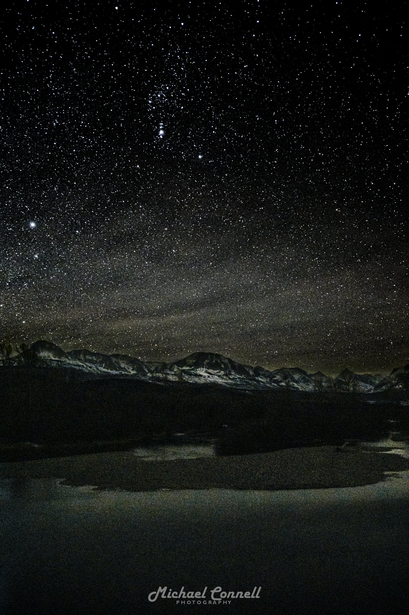 Night sky filled with countless stars above snow-capped mountain range and dark landscape, with a reflection on water in the foreground.