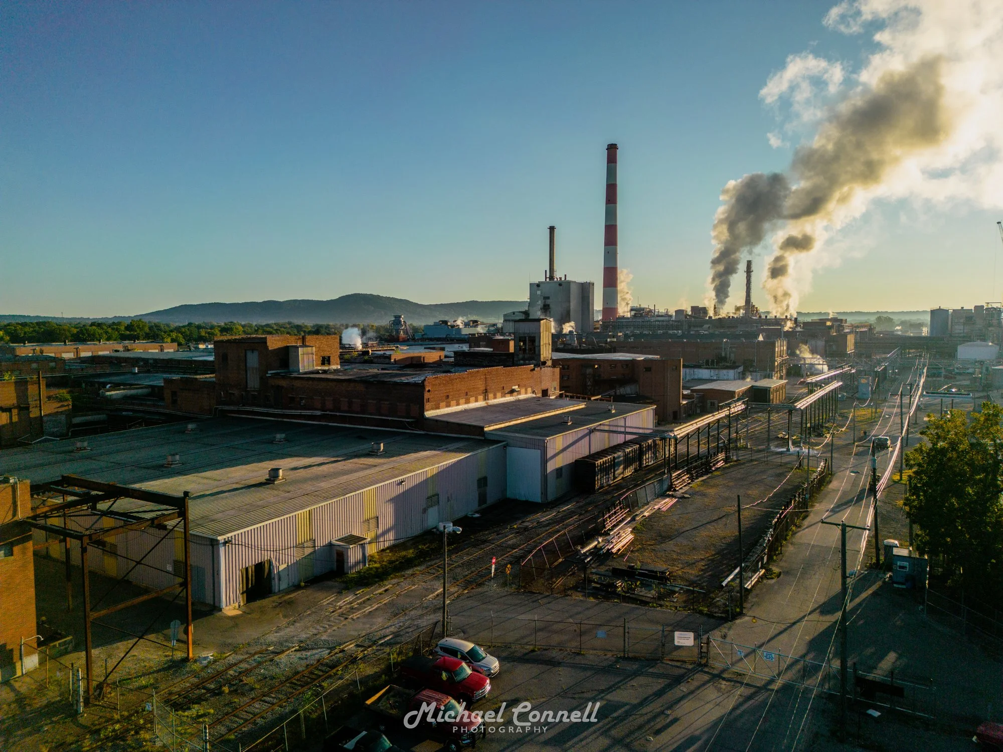 An industrial area with factories, smoke rising from chimneys, train tracks, and parked cars during sunset.