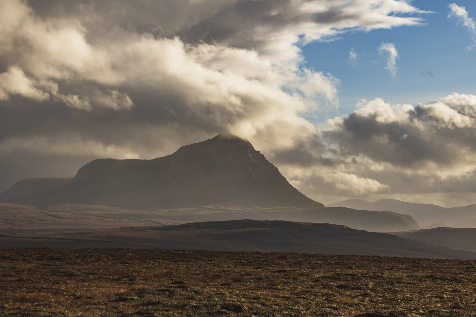 A mountain with clouds surrounding the peak, across a grassy plain with rolling hills.
