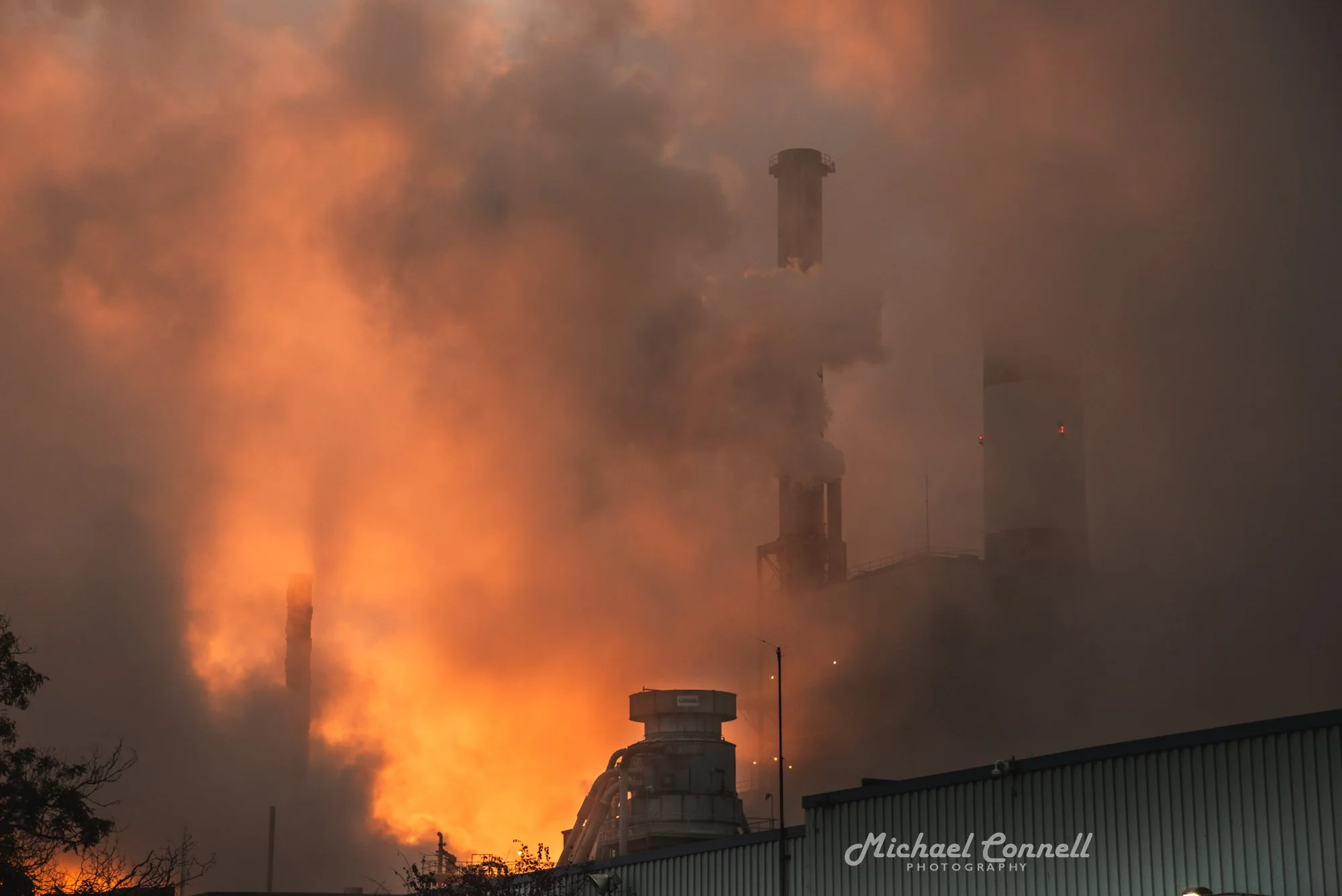 An industrial factory with smoke and fire coming out of chimneys, with orange flames and thick smoke filling the sky.