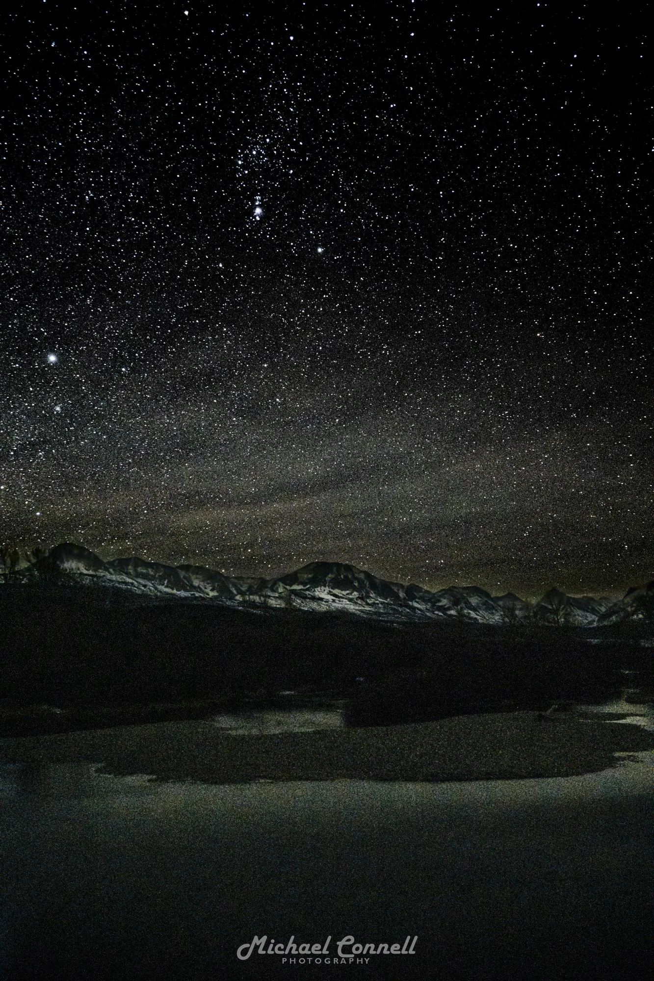Starry night sky over snow-capped mountains and a dark landscape with a body of water.