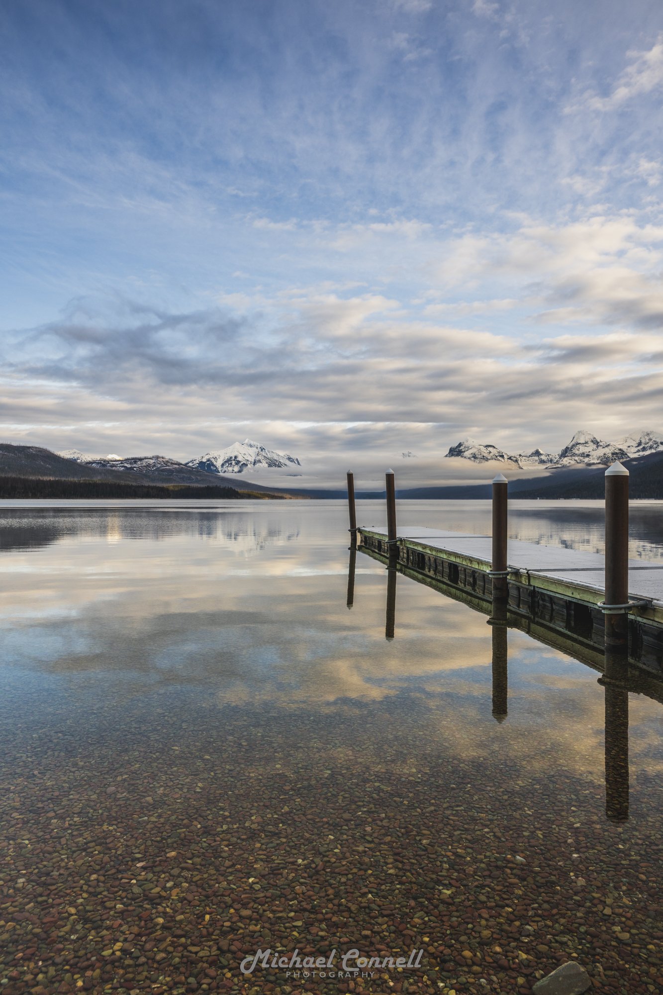 Calm lake with a dock extending into the water, snow-capped mountains in the background, cloudy sky, and their reflection in the water.