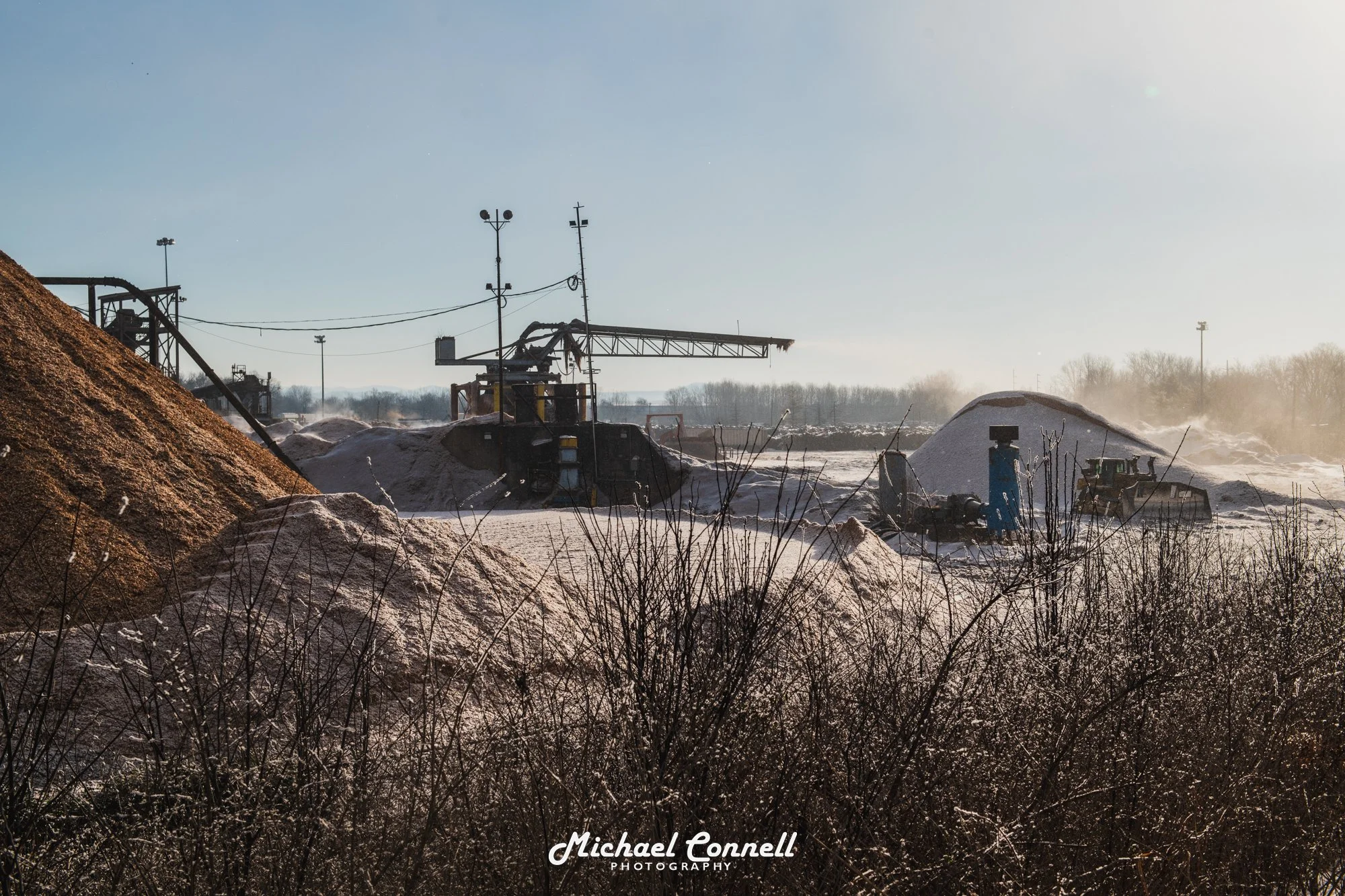 Industrial scene with piles of salt or sand, a large conveyor, and machinery in a snowy landscape with a clear sky.