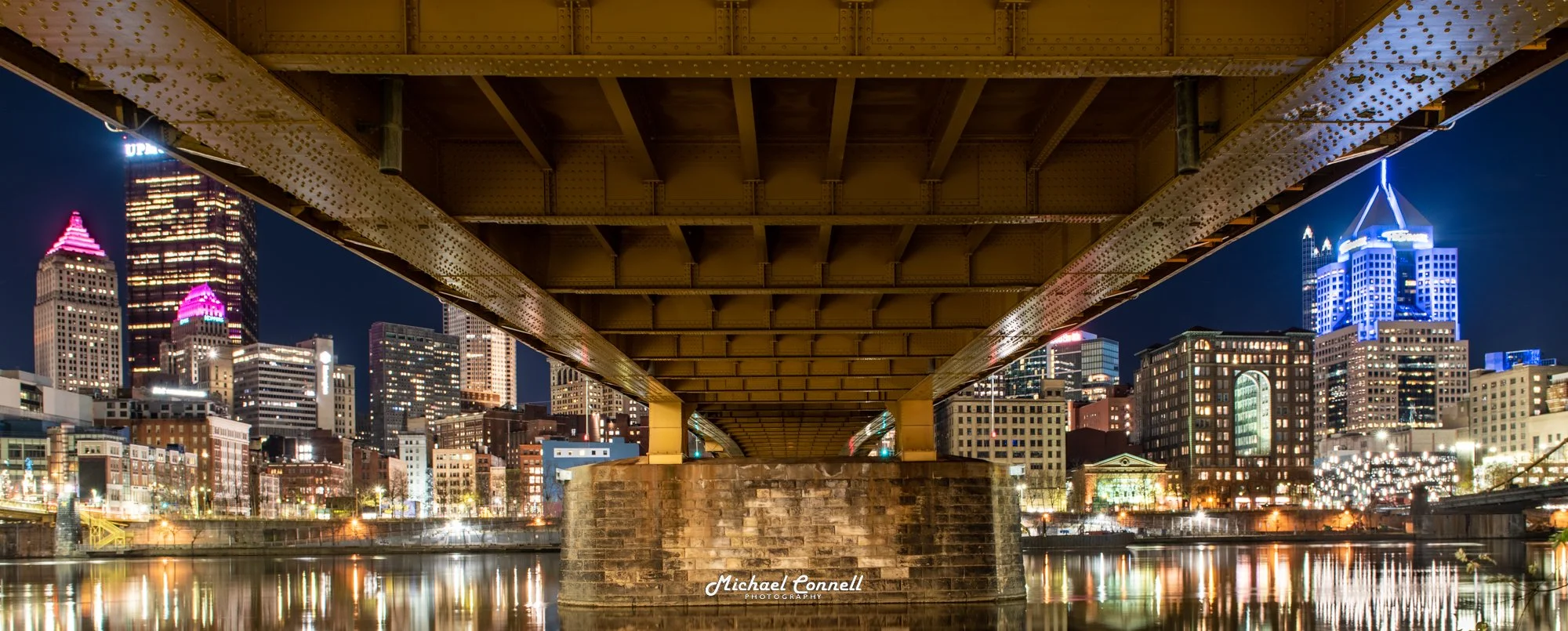 Night city skyline viewed from underneath a bridge over a river with illuminated buildings and reflections in the water.