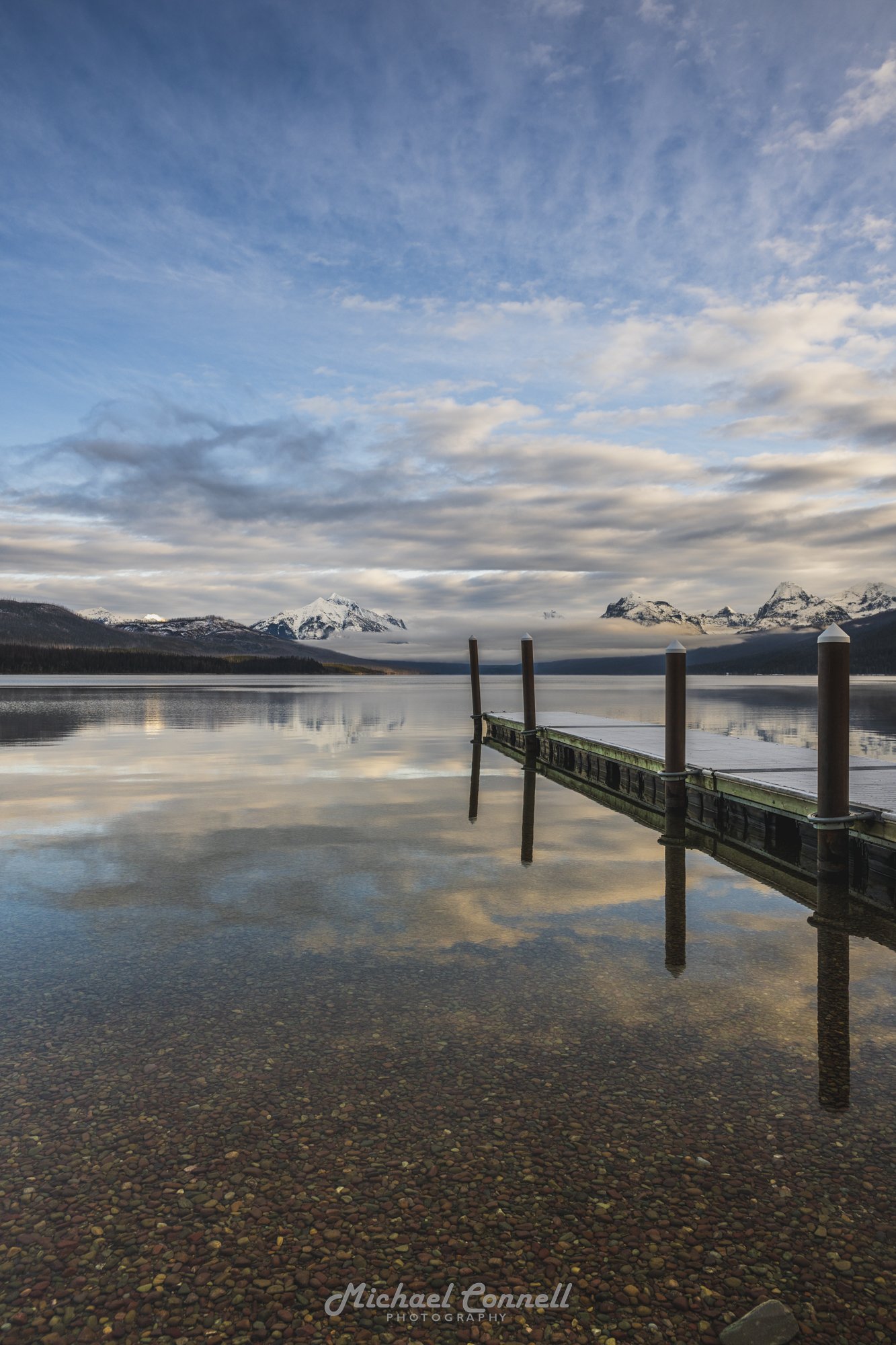 A serene mountain lake scene with calm water reflecting the sky and mountains, a wooden dock extending into the lake, snow-capped mountains in the background, and a partly cloudy sky.