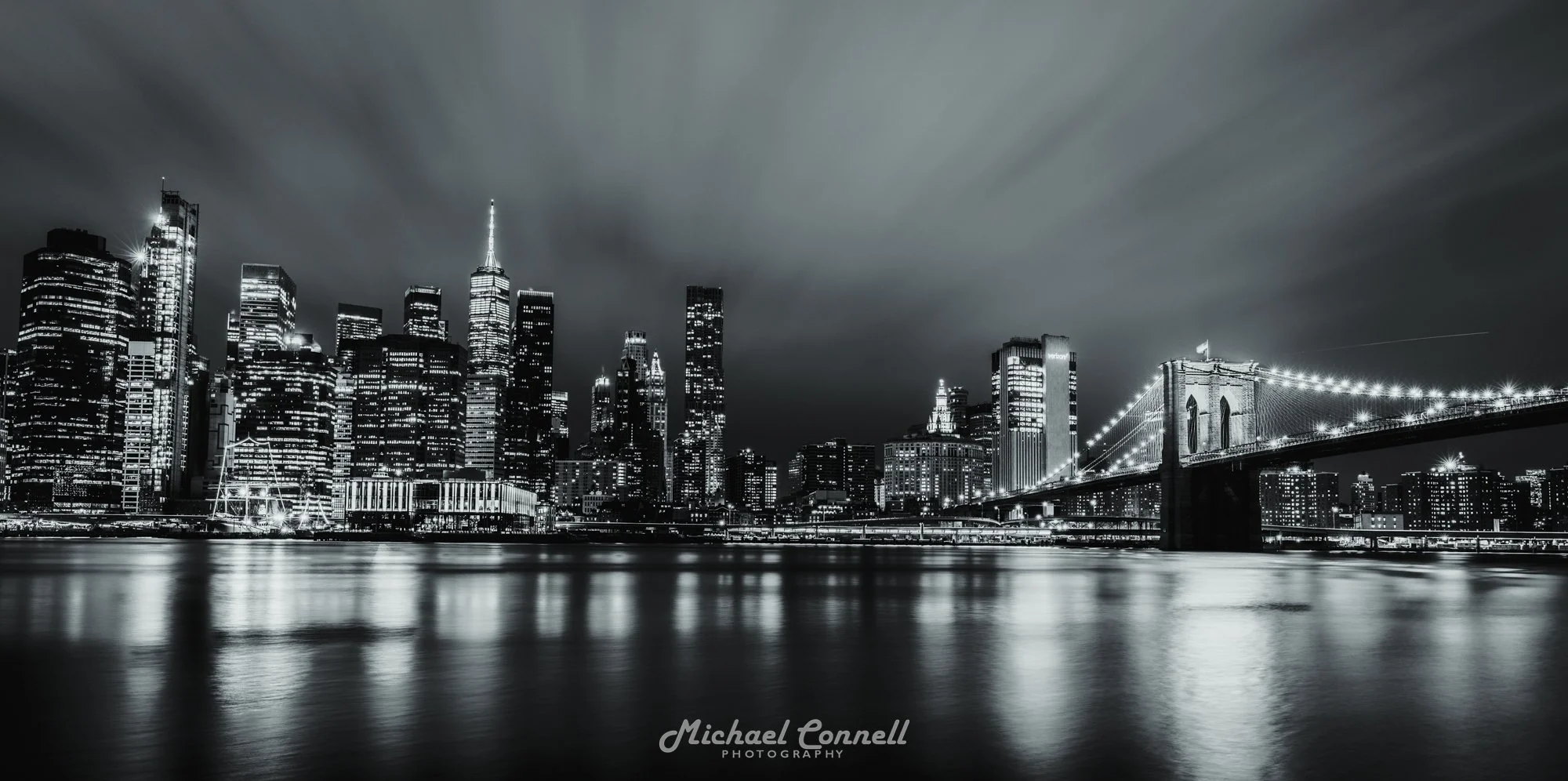 Black and white night view of New York City skyline with illuminated skyscrapers and Brooklyn Bridge over water.