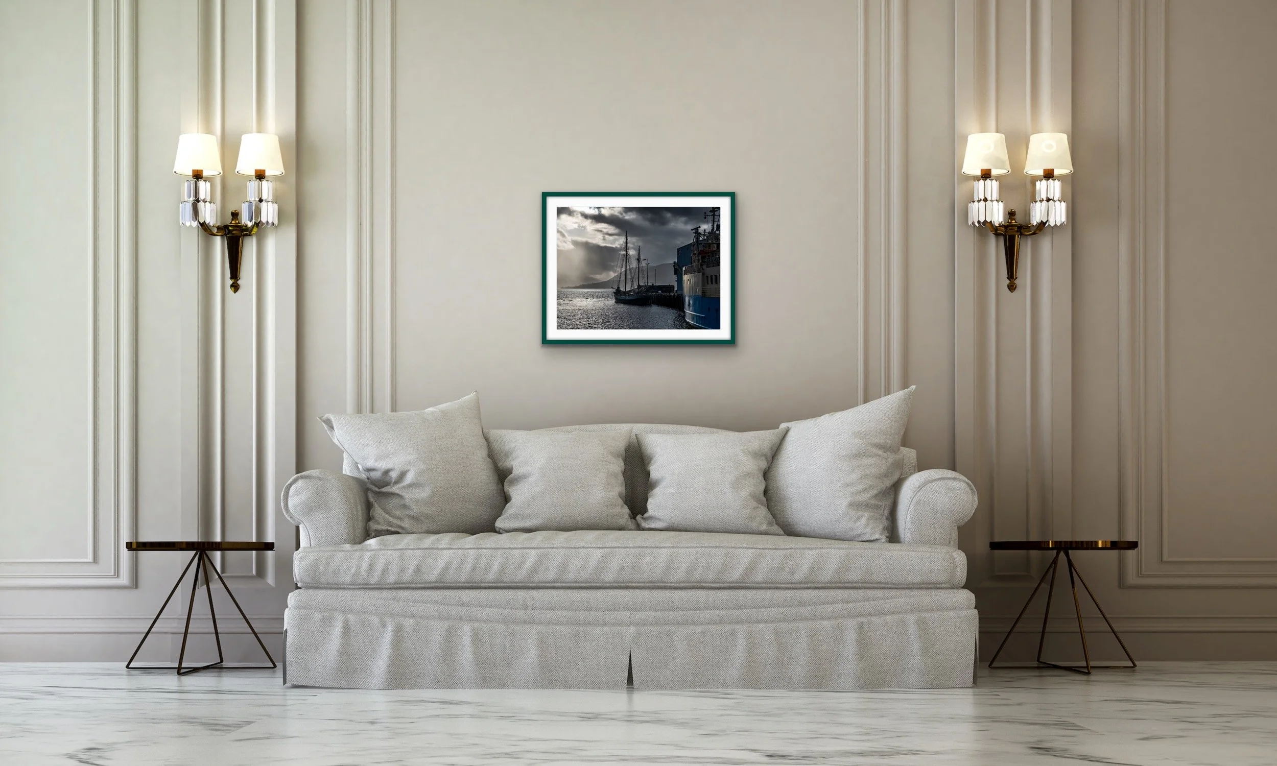 Living room with beige walls, elegant wall sconces, a light gray sofa with multiple pillows, a framed nautical photograph, and small side tables.