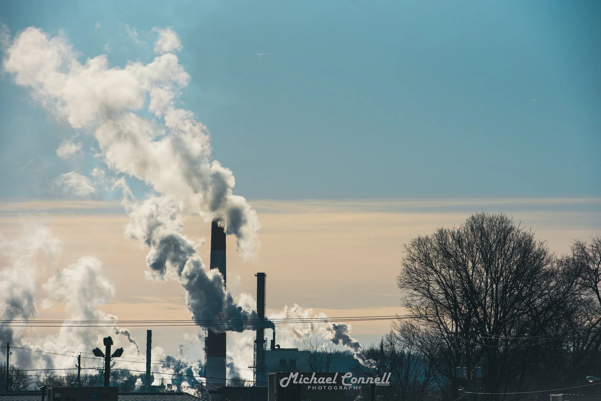 Industrial factory with tall smoke stacks emitting white smoke, and leafless trees in the foreground, under a clear blue sky.