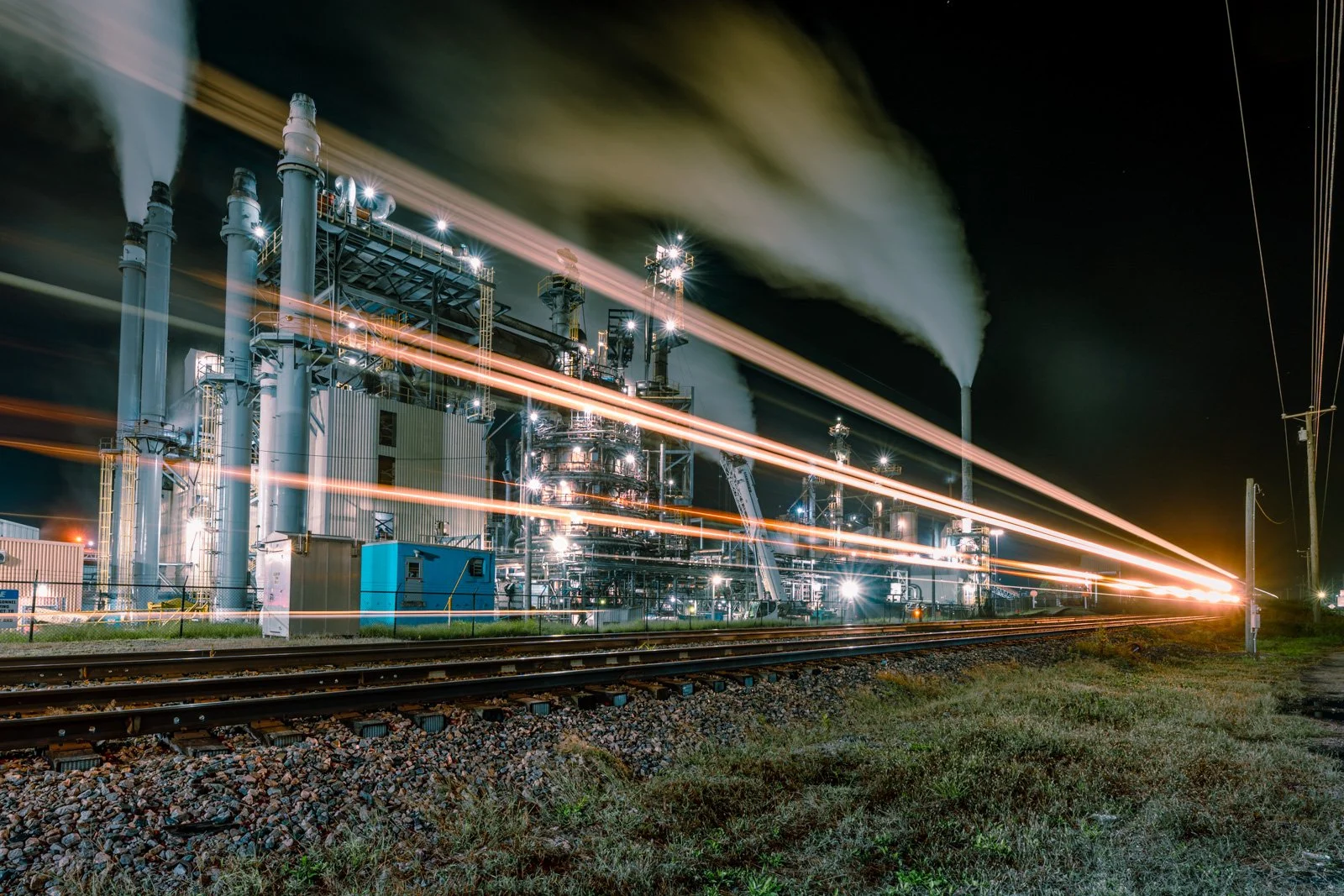 Nighttime industrial factory with smoke stacks emitting smoke, illuminated buildings, and streaks of light from moving trains or vehicles along railroad tracks in the foreground.