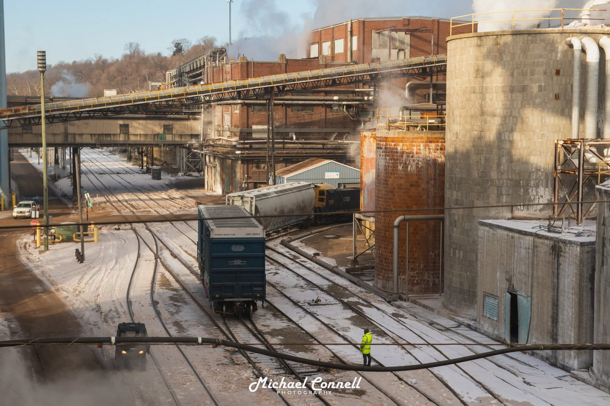 Industrial scene with train tracks, a moving freight train, large tanks, pipes, and steam emissions, with snow on the ground and a worker in a yellow safety vest.