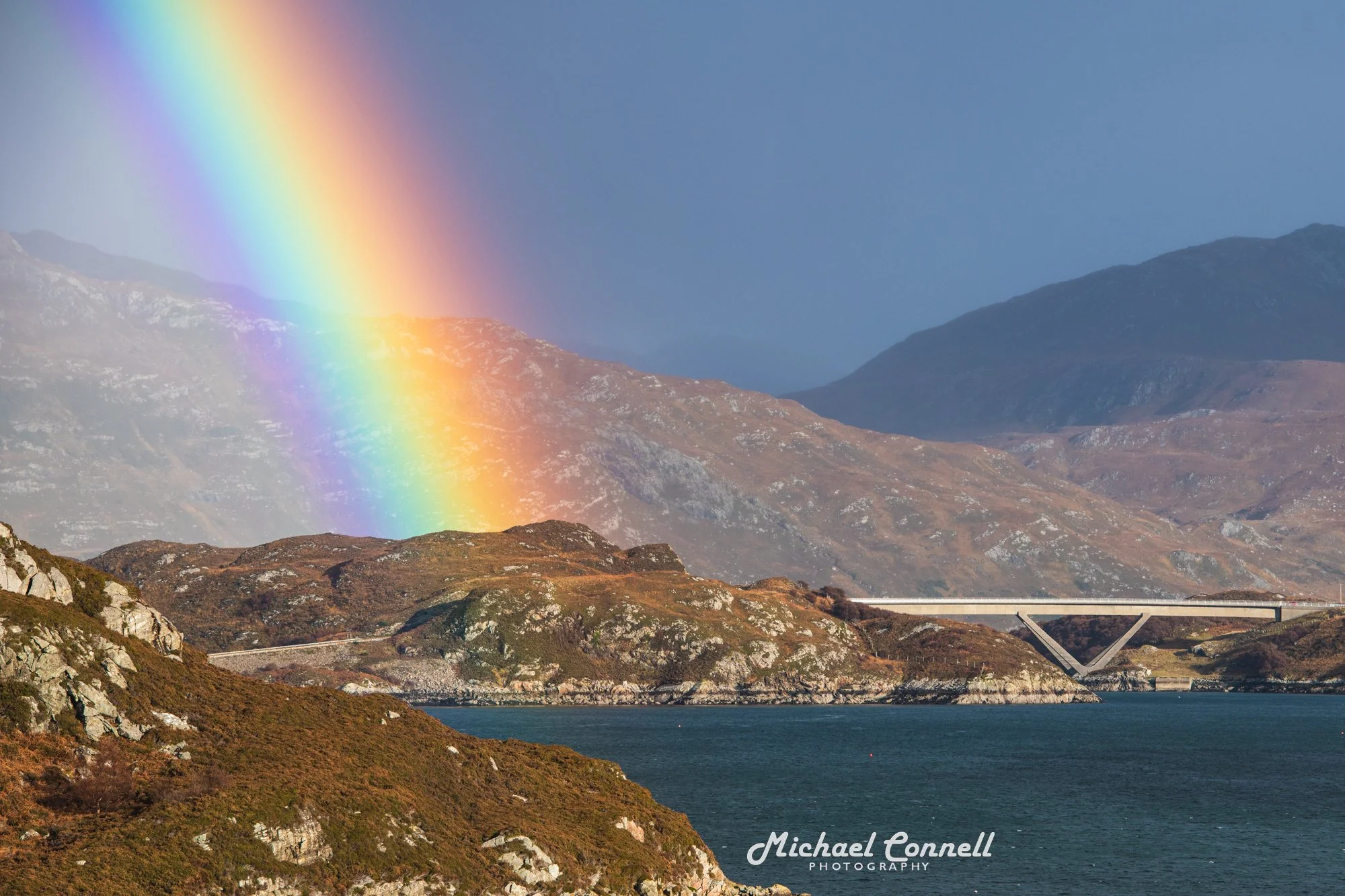 A rainbow over mountainous landscape with a body of water and a bridge in the foreground.