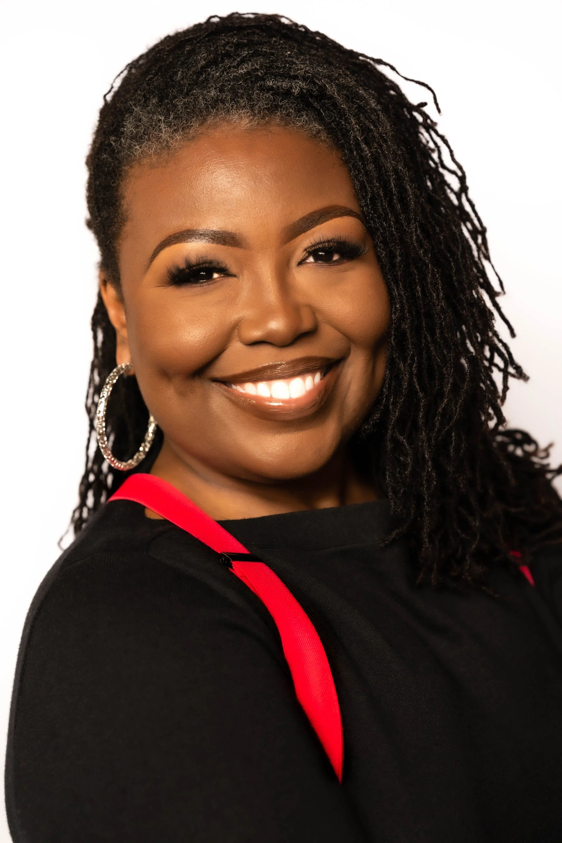 A smiling Black woman with long curly hair, wearing a black top with red accents.
