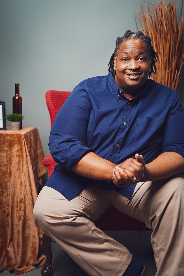 A smiling Black man wearing a blue shirt and beige pants, seated in a relaxed indoor setting.