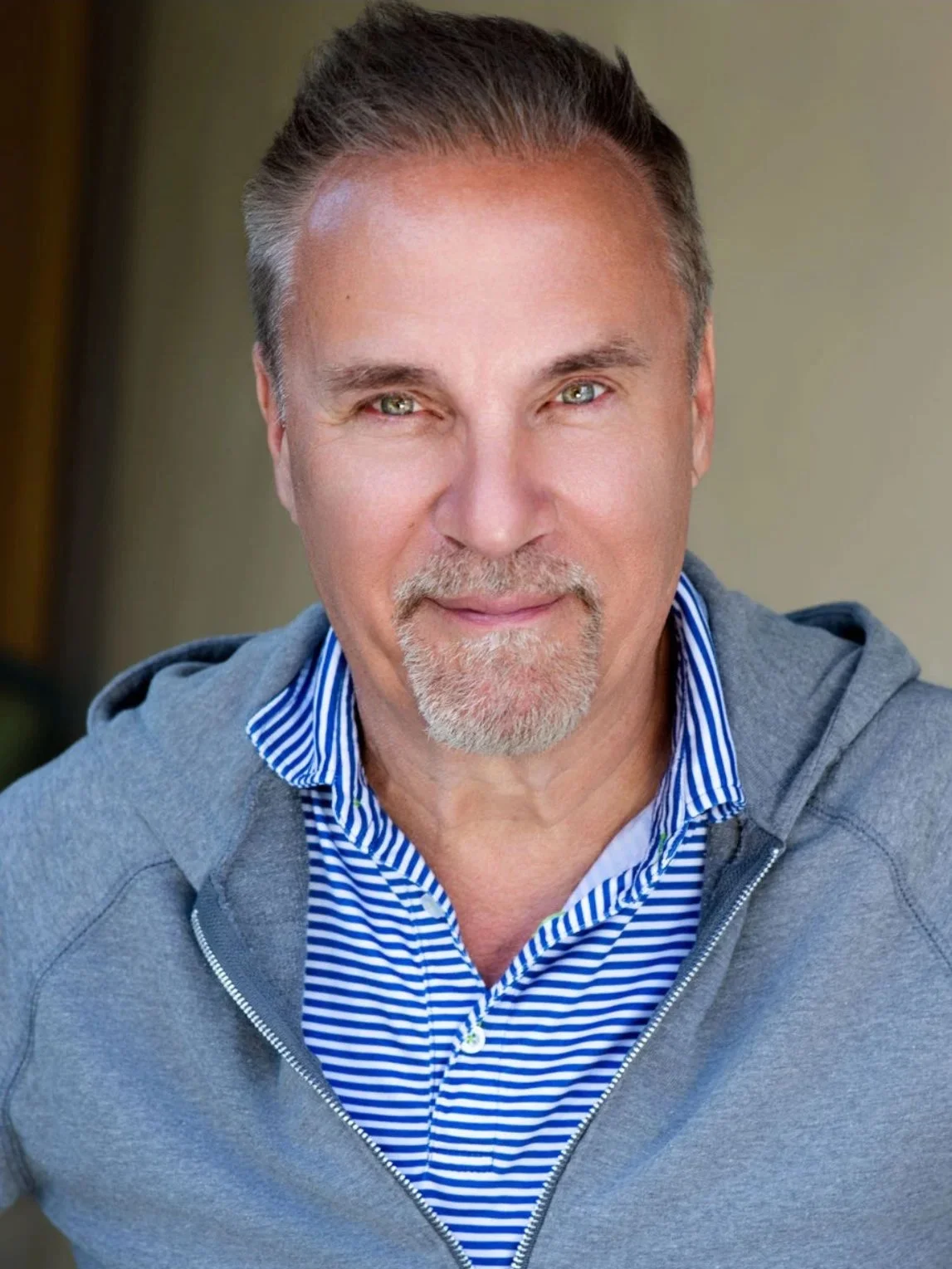 A middle-aged man with gray hair and a goatee, smiling outdoors, wearing a gray hoodie over a blue and white striped collared shirt.