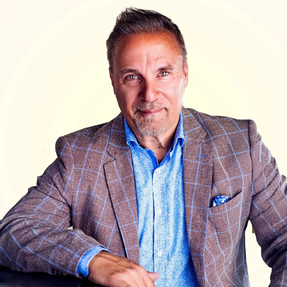 A middle-aged man with gray hair, blue eyes, and a goatee, wearing a brown checked blazer over a blue patterned shirt, posing against a light background.