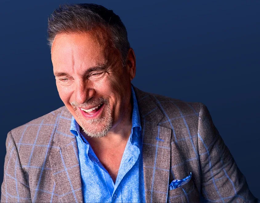 A smiling middle-aged man with short gray hair and a goatee, wearing a brown checkered blazer and a blue shirt against a plain dark background.