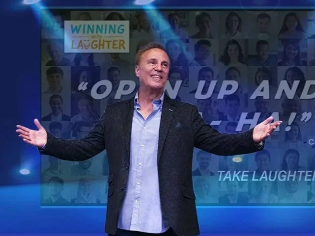 A man standing on stage with arms outstretched, smiling, with a backdrop featuring a collage of people's photos and the words, 'OPEN UP AND LAUGH,' and a sign that says, 'WINNING WITH LAUGHTER.'