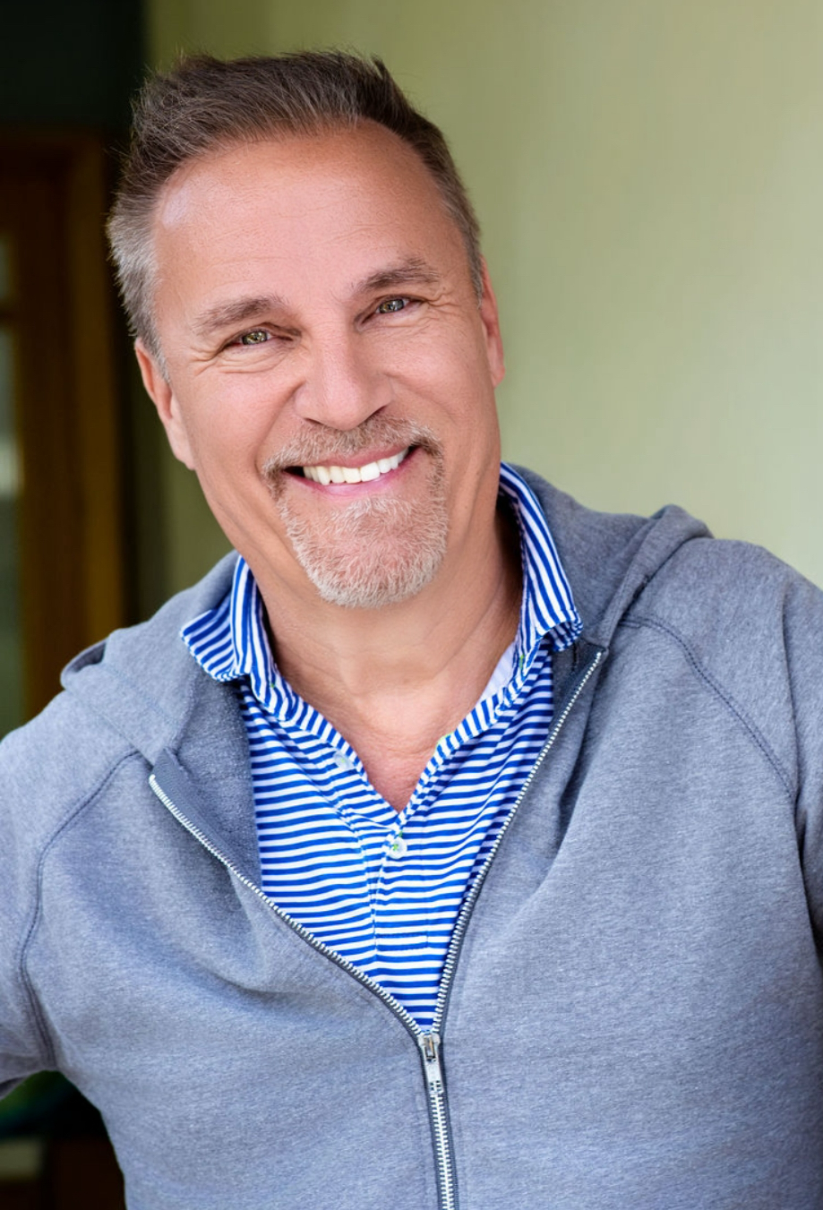 A middle-aged man with short, styled gray hair and a goatee, smiling and wearing a blue and white striped shirt with a gray zip-up hoodie