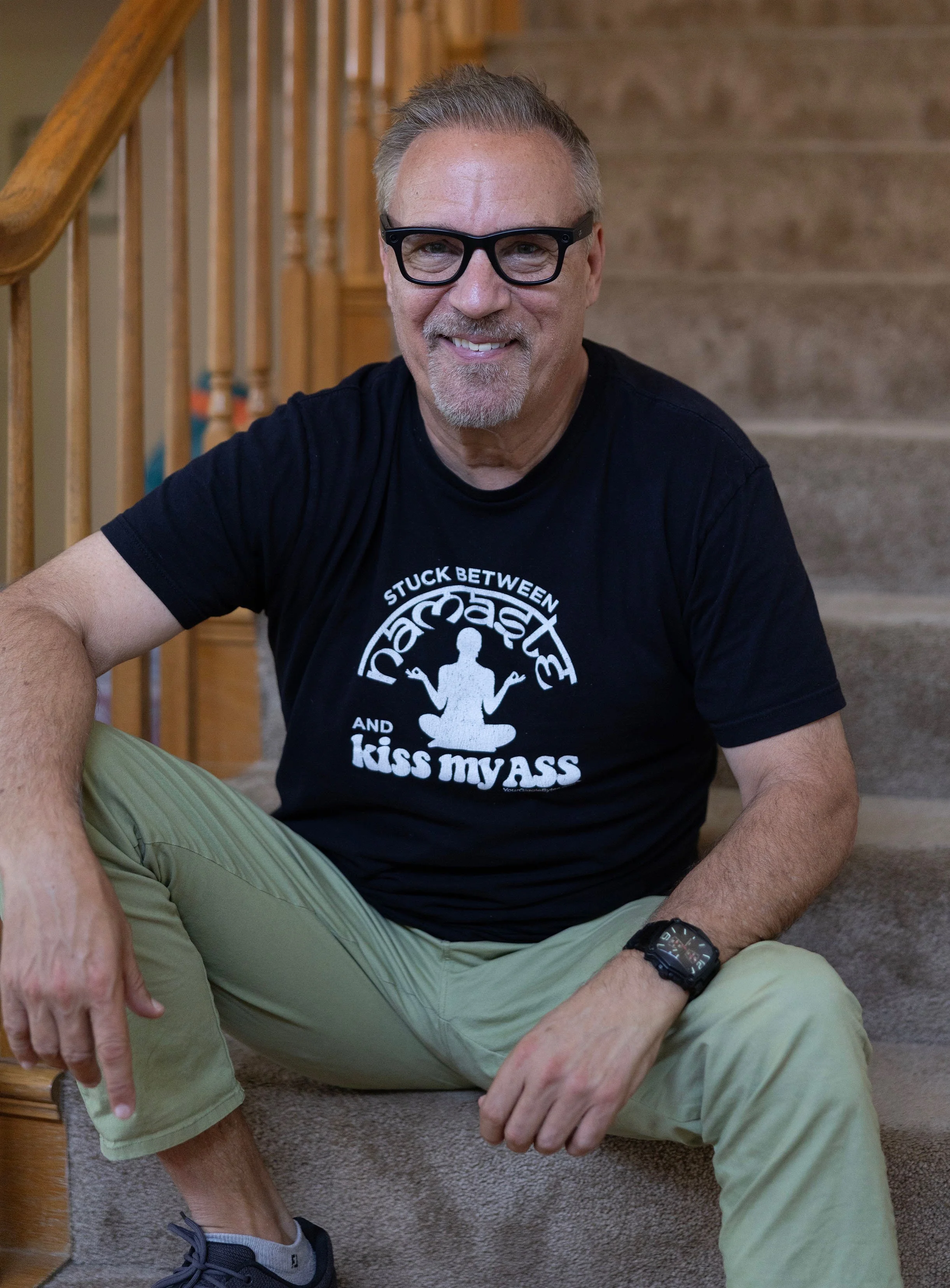 A smiling man with gray hair, glasses, and a goatee sitting on carpeted stairs. He is wearing a black T-shirt with white text and graphics, light-colored pants, a black watch, and sneakers.