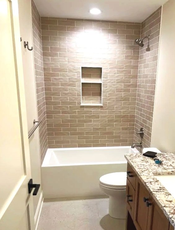 Bathroom with bathtub, toilet, granite countertop, and beige brick wall with built-in shelf