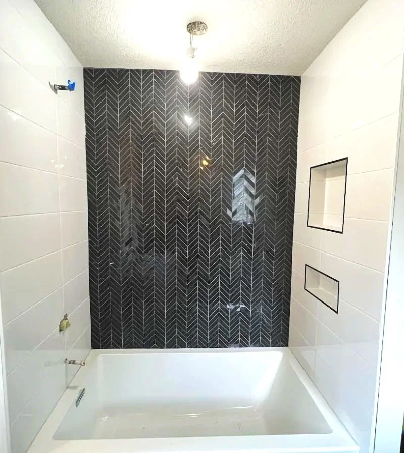 A bathroom shower with a black tiled accent wall featuring a herringbone pattern, white tile walls, and built-in niche shelves.