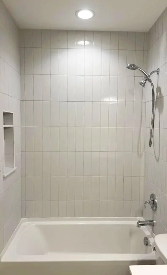 Bathroom with white tiled walls, bathtub, showerhead mounted on the wall, built-in shower niche, and a ceiling light.