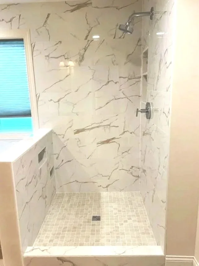 A walk-in shower with beige marble walls and beige mosaic tile floor, a chrome showerhead, and a window with blinds.