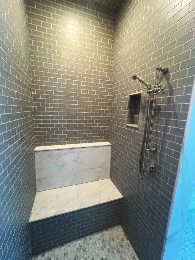 A modern shower area with dark gray subway tile walls, a built-in marble bench, a small wall niche, and a rain shower head with a handheld shower attachment.