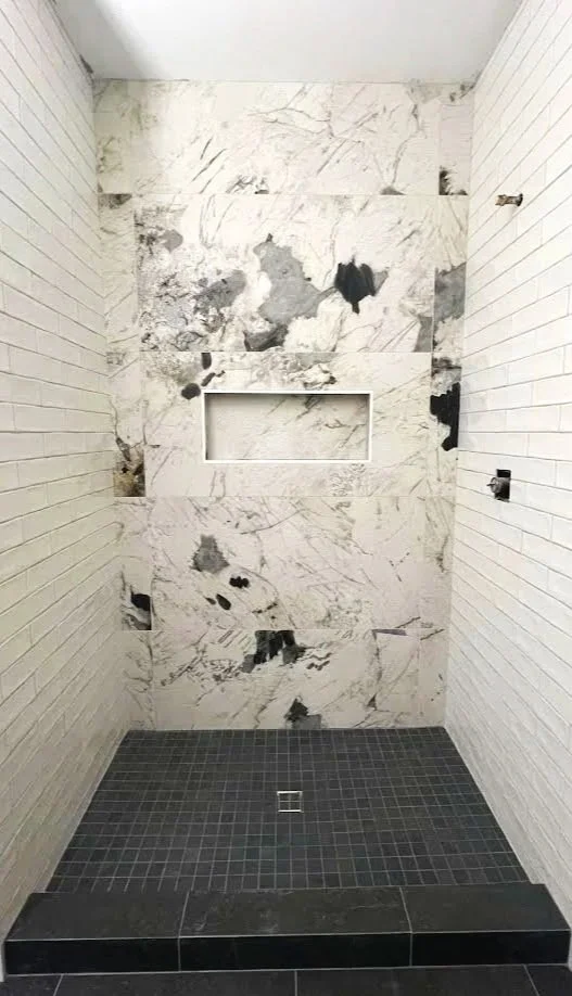 A partially completed shower with marble tiling on the back wall, some tiles missing, and black small square floor tiles with a drain in the center.