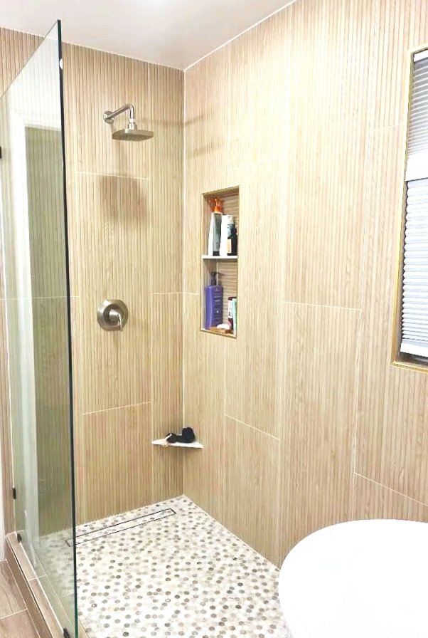A modern shower area with beige tiled walls, a built-in shelf with toiletries, and a glass shower enclosure. A window with blinds is visible on the right.